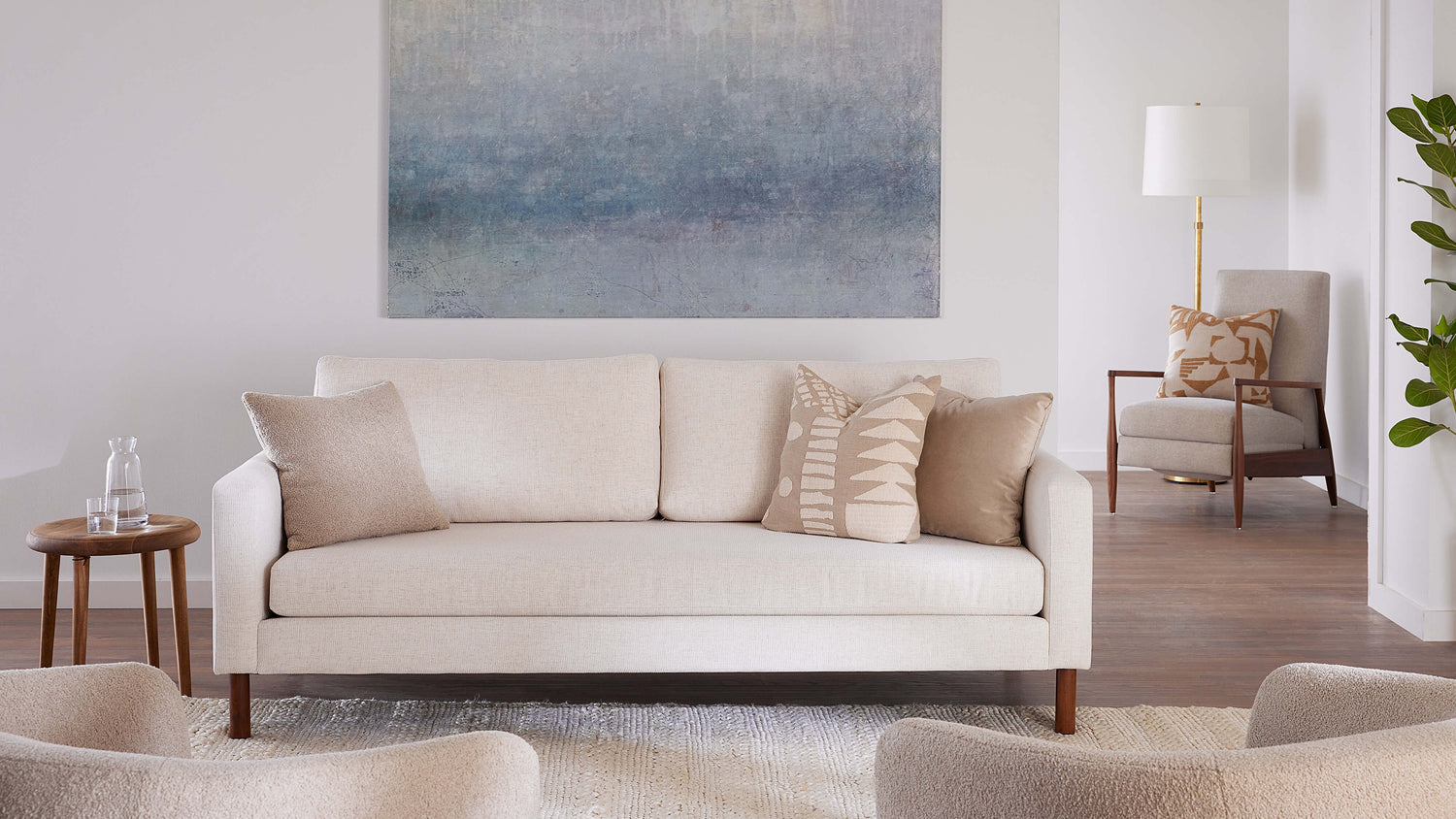 A minimalistic living room features a beige sofa with neutral-toned pillows, a round wooden side table with water glasses, and an abstract wall painting. In the background, a beige armchair, a floor lamp, and a leafy plant add to the serene atmosphere.