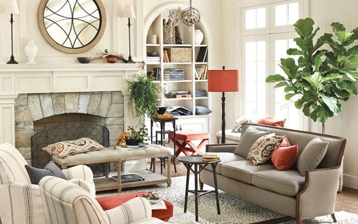 A cozy living room featuring a neutral-toned sofa with red and patterned pillows, armchairs, a stone fireplace, and a round mirror above the mantel. A tall potted plant, assorted books and decor on shelves, and a red floor lamp add warmth and character to the space.