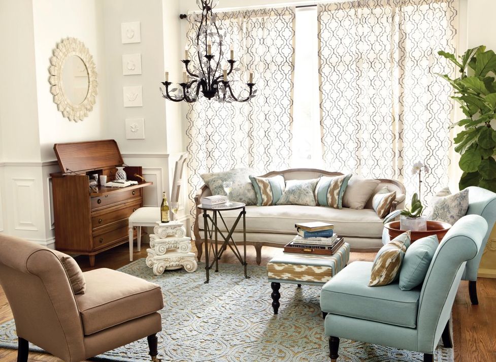 A cozy living room features a beige sofa, patterned armchairs, and a blue chaise lounge. A wooden cabinet, coffee table, and side tables are decorated with books and a plant. The room is well-lit with natural light through large, patterned curtains. A chandelier hangs overhead.