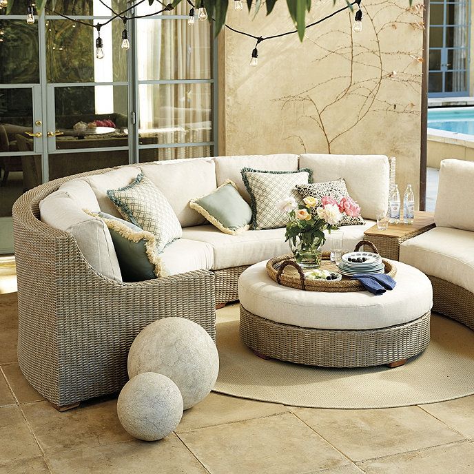 A cozy outdoor seating area features a beige wicker curved sofa with cushions and pillows, surrounding a matching round ottoman with a tray of flowers and drinks. Two large decorative stone orbs sit on the tiled floor. String lights hang overhead, and a pool is visible in the background.