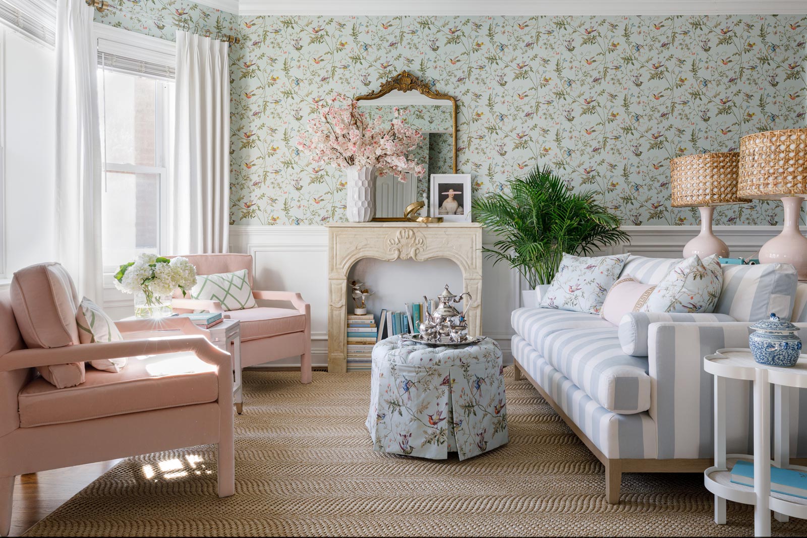 A cozy living room features pastel-colored furniture and floral-themed decor. Two pink armchairs flank a floral ottoman in front of a striped sofa. A fireplace with a mirror above is adorned with flowers, and a large plant sits beside a side table with a blue vase.