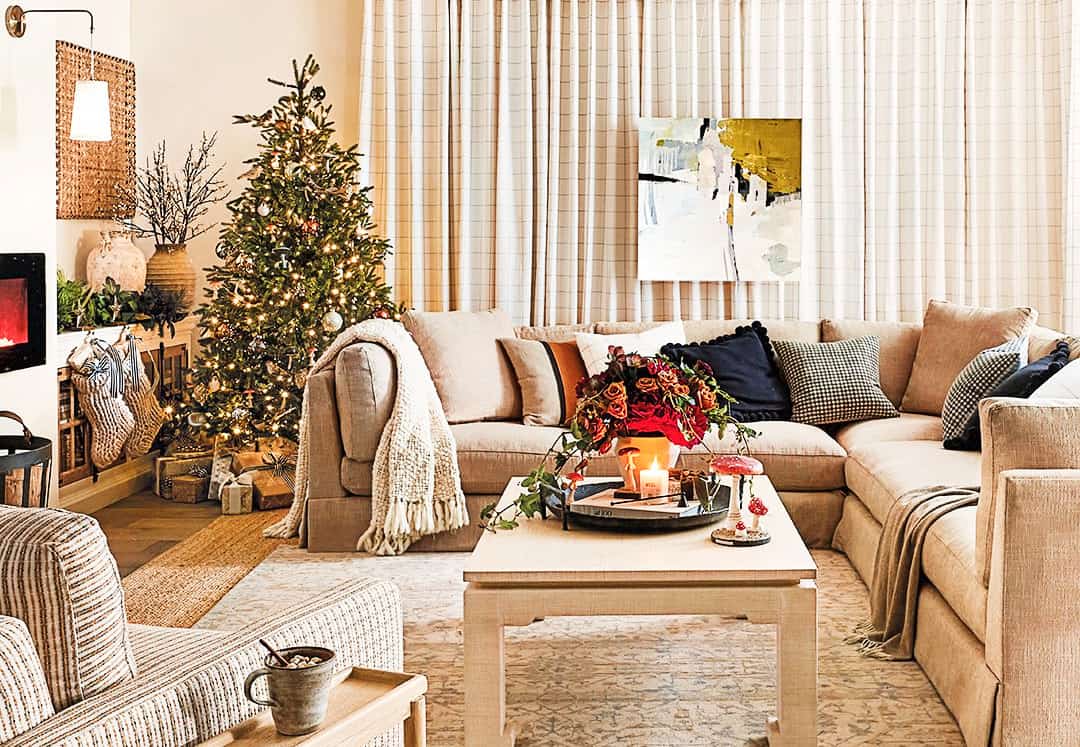 A cozy living room features a large beige sectional sofa adorned with pillows and throws. A decorated Christmas tree stands next to a window with long curtains, while a coffee table in front displays a floral centerpiece and candles. A painting hangs on the wall.