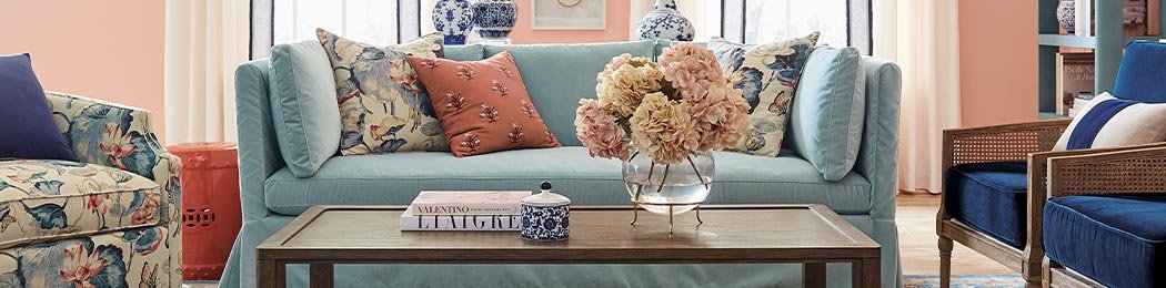 A stylish living room featuring a light blue sofa adorned with patterned cushions and a coral throw pillow. In front of the sofa is a wooden coffee table with books, a decorative jar, and a vase of flowers. Blue armchairs and floral elements complete the decor.