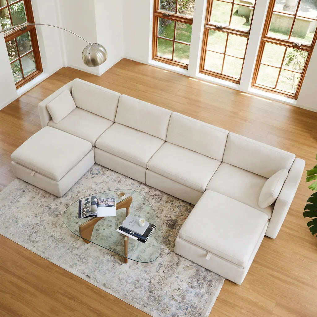 A modern living room with a beige L-shaped sectional sofa, a glass coffee table on a patterned area rug, and large windows offering plenty of natural light. The sofa has matching ottomans and a sleek, curved floor lamp stands beside it. Wooden floors and a green plant near the sofa add to the decor.