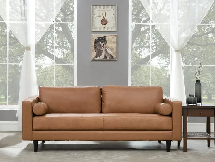 A stylish living room with a brown leather sofa centered against a gray wall. Two framed art pieces hang above the sofa. Light-filled large windows with white curtains offer a view of lush green trees. A wooden side table with decorative items stands to the right.