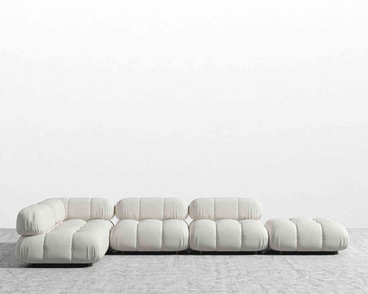 The Belia Modular Sectional, featuring white, tufted cushions, is arranged in an L-shape against a plain white wall on the gray carpeted floor. This sofa design showcases low-profile seating and a minimalist aesthetic.