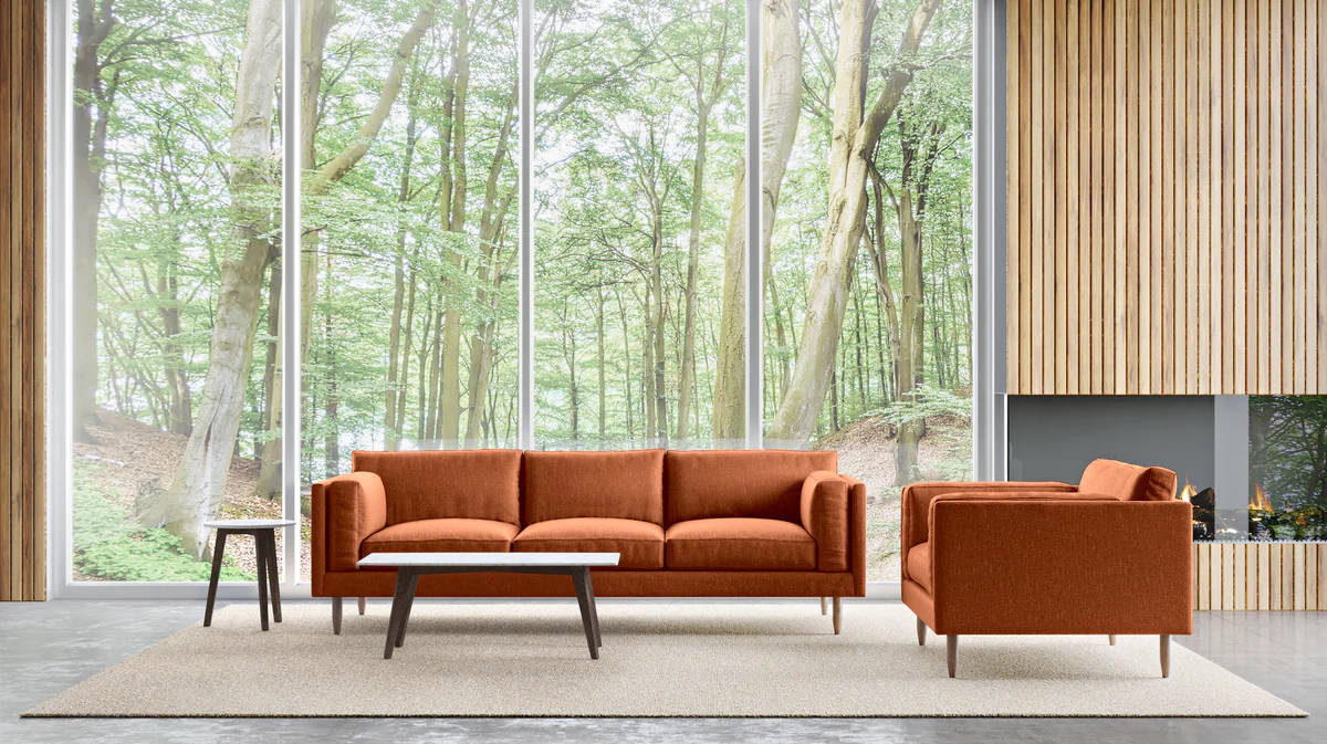 A modern living room with floor-to-ceiling windows showcasing a forest view. The space features an orange sofa, matching armchair, wooden coffee table, a small side table, and a minimalist fireplace. Light wooden paneling decorates one wall, adding warmth to the room.