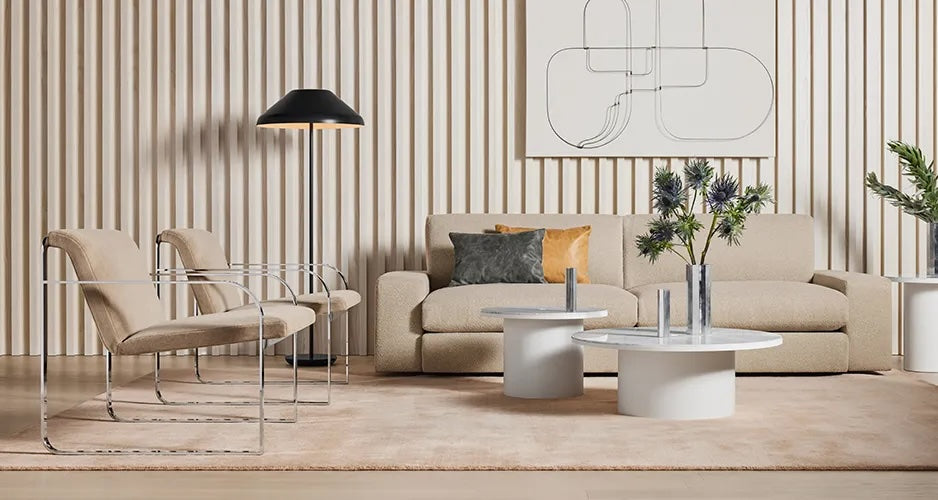 A modern living room featuring a beige sofa with two cushions, two beige chairs with metal frames, and two round white coffee tables with vases. A black floor lamp stands in the corner, and abstract wall art hangs above the sofa. The wall has vertical wooden panels.