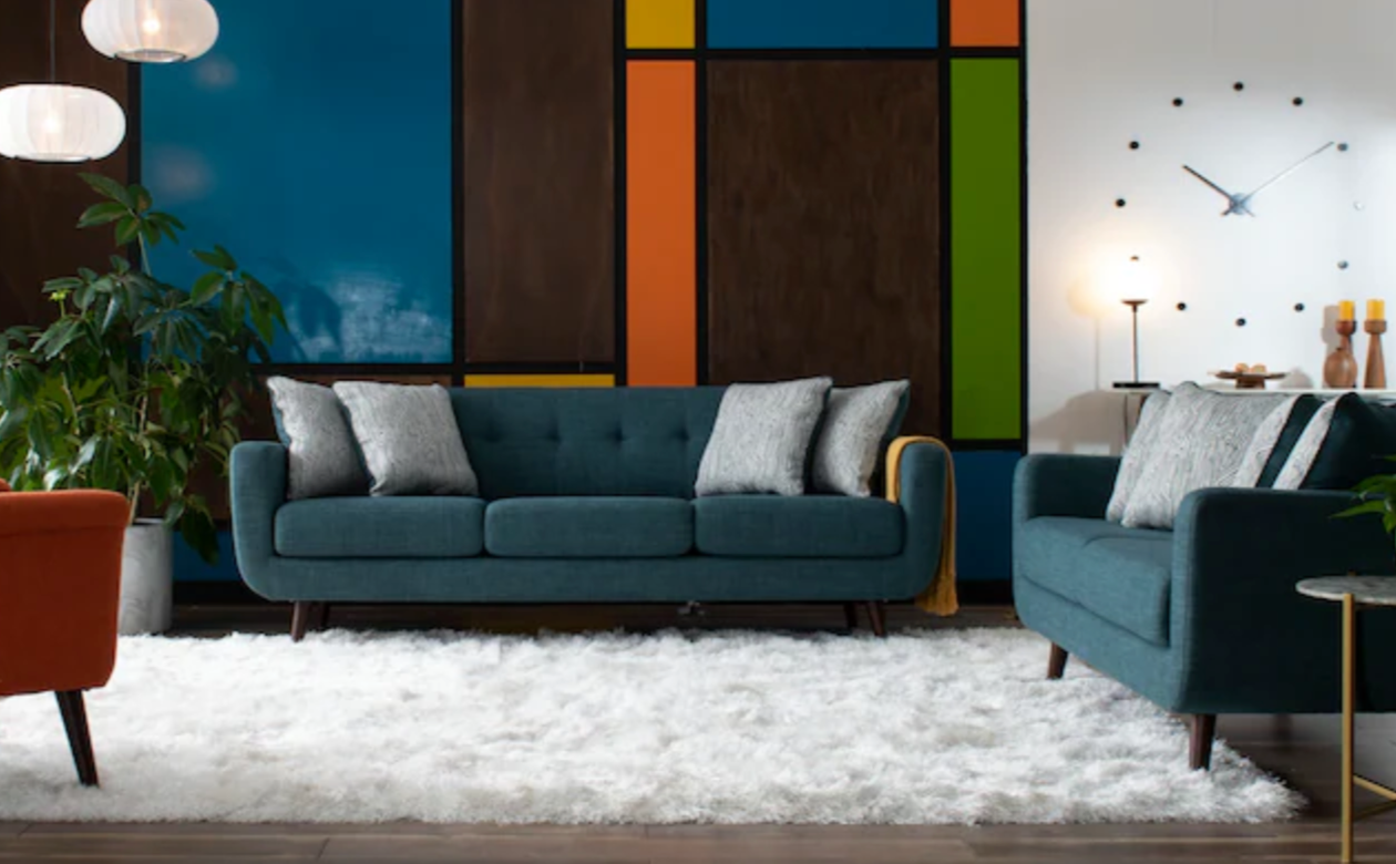 A modern living room with a dark blue sofa and matching armchair on a fluffy white rug. The wall behind features a colorful, geometric design with blue, orange, and green panels. The room is decorated with plants, a clock, and minimalist lighting.