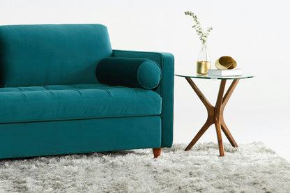 A Briar Sleeper Sofa in teal, featuring a cylindrical cushion, rests on a fluffy light gray rug. Beside it is a wooden side table with a glass top, adorned with a small vase containing greenery and a decorative gold object.