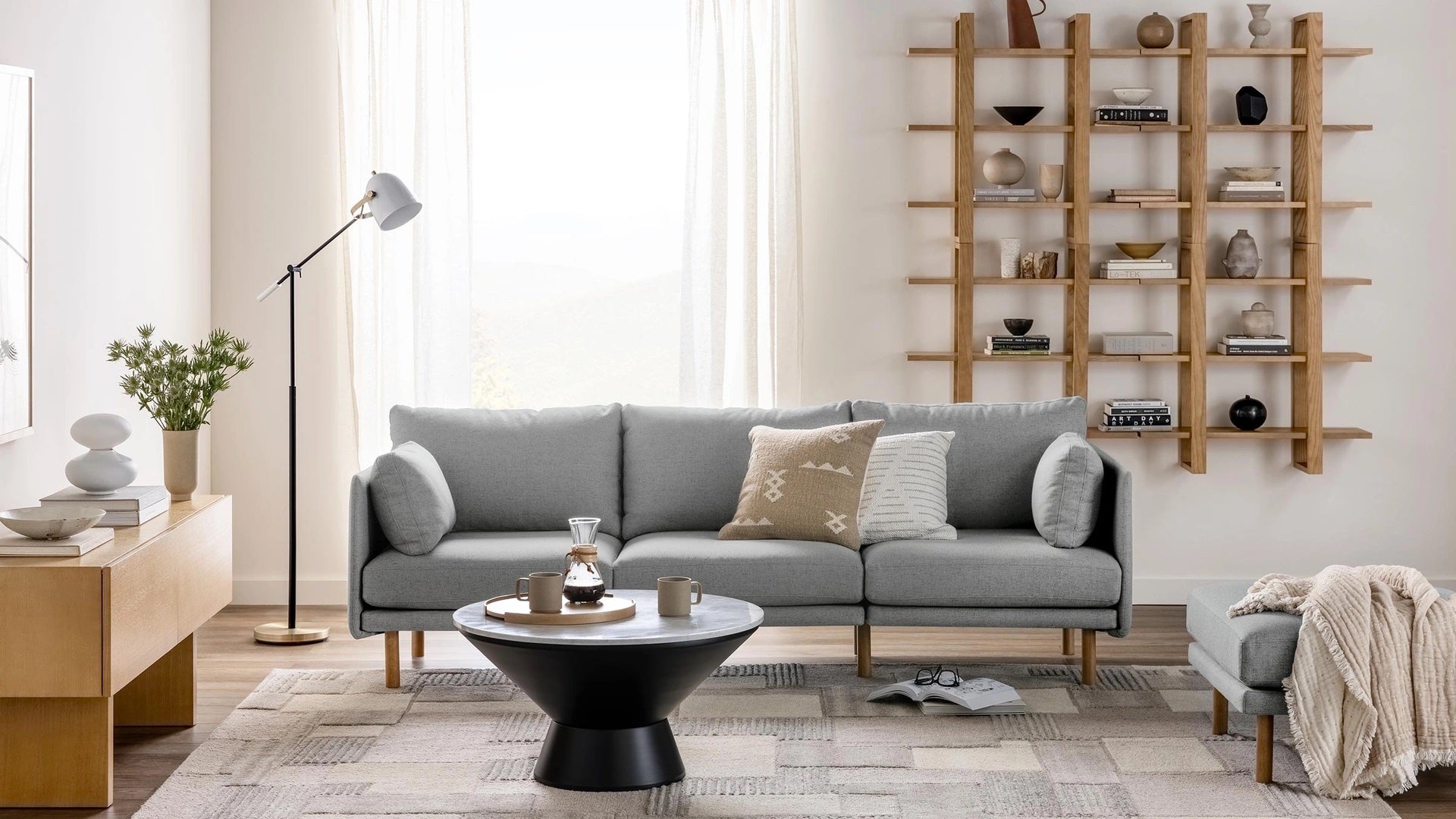 A modern living room with a light gray sofa adorned with patterned throw pillows, a round black coffee table, and a light wooden floor lamp. Shelves filled with decor items are mounted on the wall behind the sofa. A large window with sheer curtains is in the background.