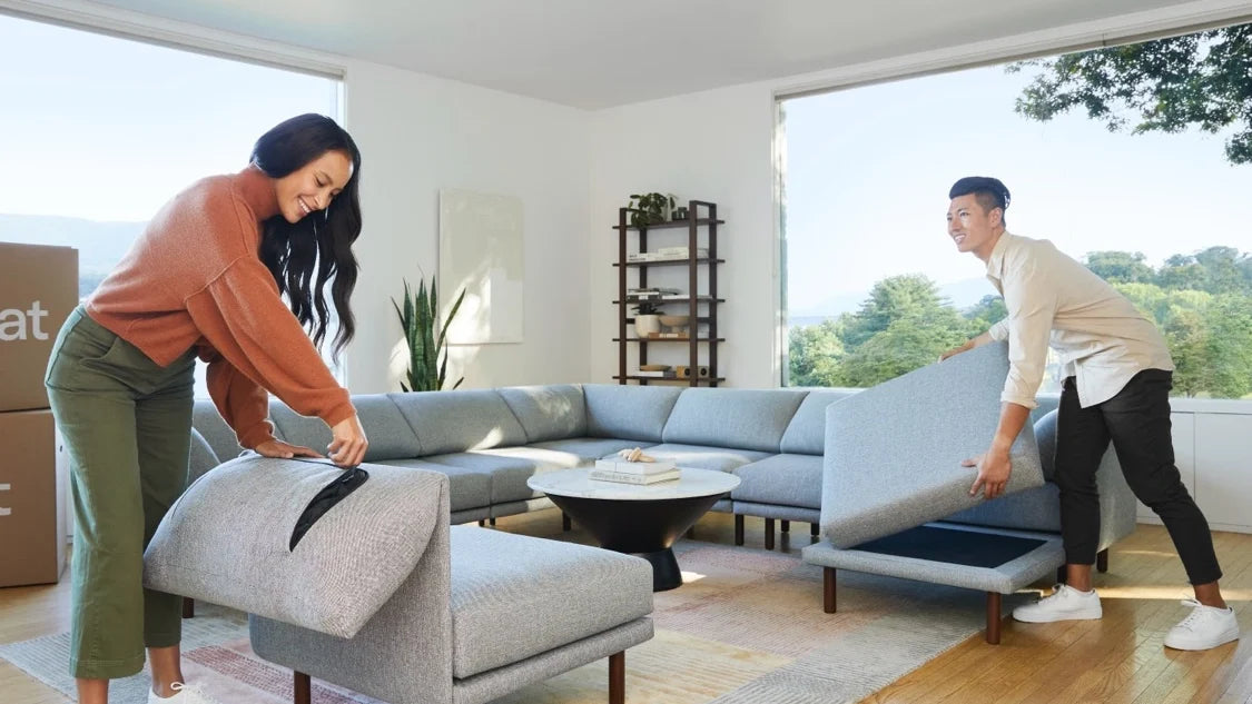 A woman and a man, both smiling, are assembling a sectional sofa in a bright living room with large windows. The woman is attaching a cushion, while the man lifts another part of the sectional. There are moving boxes in the background.