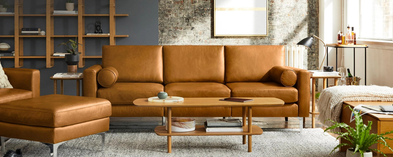 A modern living room featuring a brown leather sofa and matching ottoman. A wooden coffee table with decor items sits in front of the sofa. A textured brick wall, bookshelves, framed artwork, and a floor lamp complete the decor with natural light coming through a window.