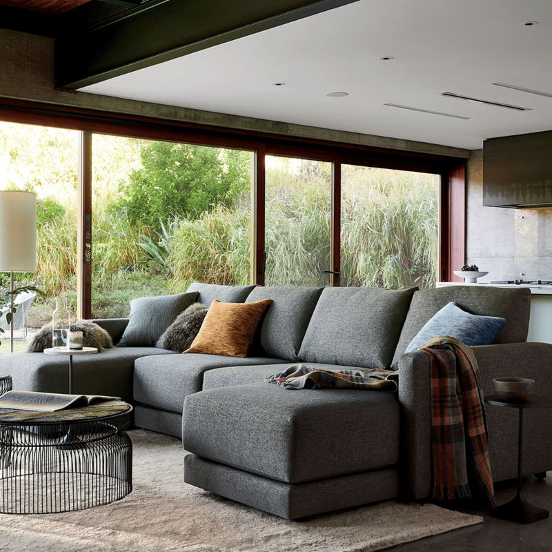 A modern living room with a large gray sectional sofa adorned with pillows and a blanket. A round black coffee table, floor lamp, and large windows with views of greenery complete the cozy space. A flat-screen TV is mounted on the wall in the background.