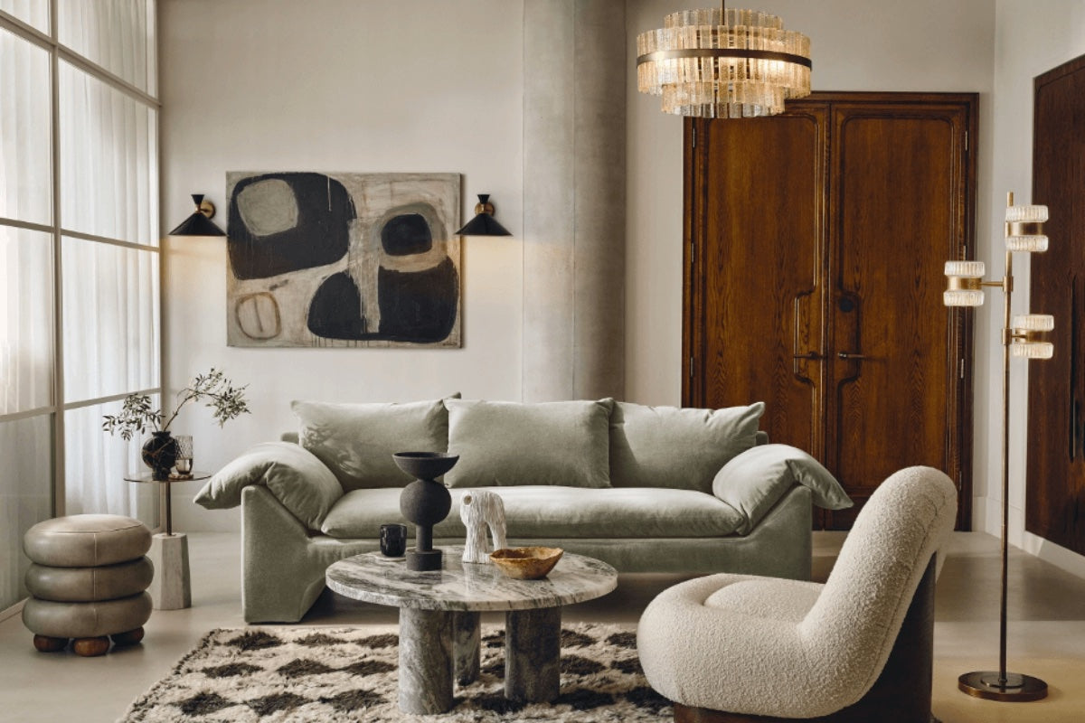 A modern living room with a plush, light gray sofa, a round marble coffee table, and a cream-colored lounge chair on a geometric-patterned rug. A large abstract painting, elegant floor lamp, decorative items, and wooden double doors adorn the space.