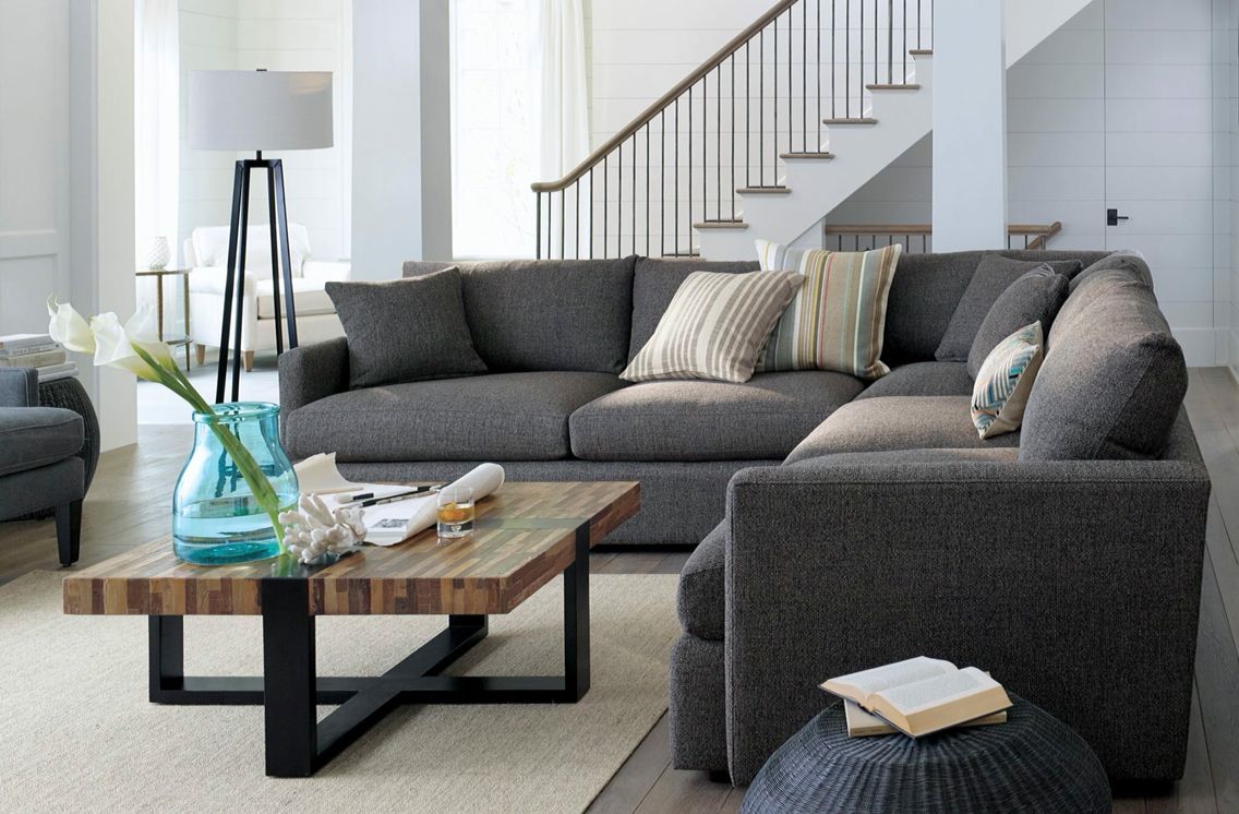 A modern living room features a large, gray sectional sofa adorned with striped and patterned cushions. A rustic wooden coffee table displays a blue glass vase with flowers, books, and a decorative shell. A staircase with a wooden railing is in the background.