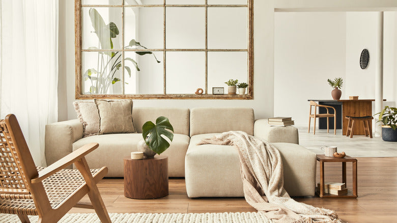 A modern living room features a beige sectional sofa with a blanket draped over it and a potted plant on a wooden side table. A wicker chair sits nearby. A large window with a wooden frame separates the dining area in the background, which has a wooden table and chairs.