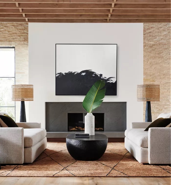 A modern living room with two beige sofas facing a black round coffee table. The table holds a vase with a large green leaf. Behind the table, a fireplace is centered in a white wall, above which hangs abstract black and white art. Two lamps flank the fireplace.