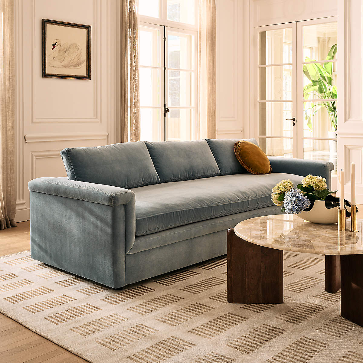 A stylish living room showcases the Ceva 103" Light Blue Performance Velvet Sofa, elegantly adorned with a yellow cushion. A patterned cream rug adds texture beneath a round wooden coffee table, topped with marble and holding a vase of flowers. Large windows and framed art pieces further enhance the ambiance.