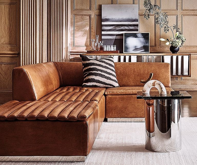 A cozy living room features a brown leather sectional sofa with a striped cushion, a black glass-topped metal side table with decorative items, and a wood-paneled wall adorned with monochrome artwork. A minimalist console and a potted plant complete the decor.