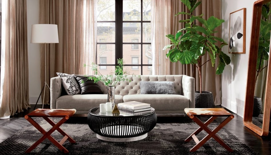 A modern living room with a beige sofa adorned with pillows, a round black coffee table topped with decor items, and two wooden stools. The room features large windows with beige curtains, a floor lamp, a tall plant, and a large mirror leaning against the wall.