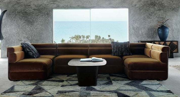 A modern living room features a large, brown L-shaped sofa with three patterned pillows. A wooden coffee table sits on a geometric-patterned rug. Large windows in the background offer a view of the ocean and palm trees. Decor includes a tall vase and a sideboard.