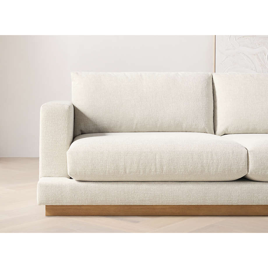 A close-up view showcases the Tidal Apartment 83" Sofa, designed with modern beige textured fabric, wide cushions, and a wooden base. The background features a light-colored wall with minimalist decor elements.