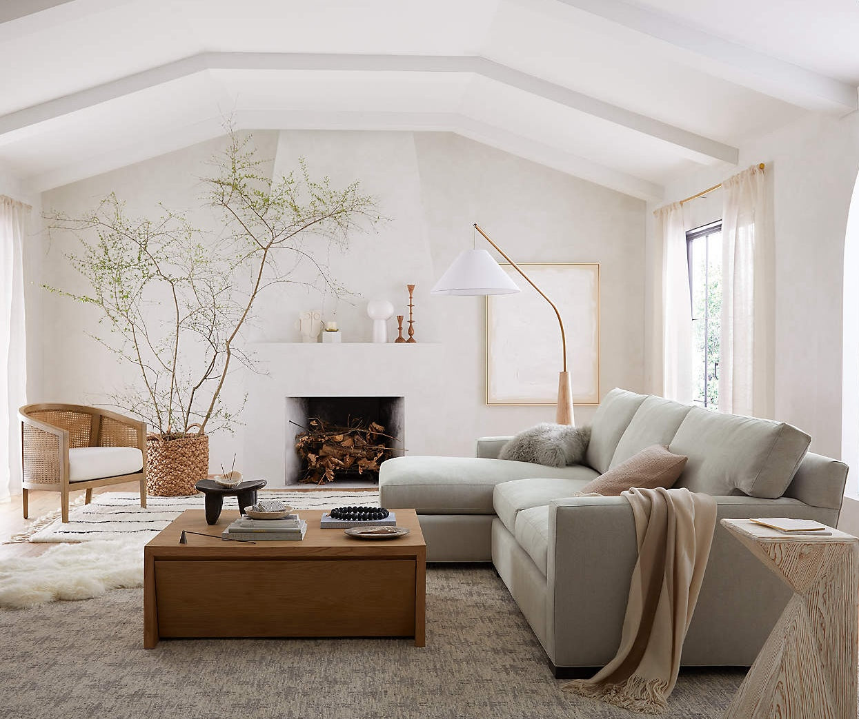 A bright, minimalist living room with a vaulted ceiling features a light gray sectional sofa, a wooden coffee table, and a fireplace. A slender floor lamp and a tall potted plant add decorative touches. Large windows with light curtains allow natural light to fill the room.