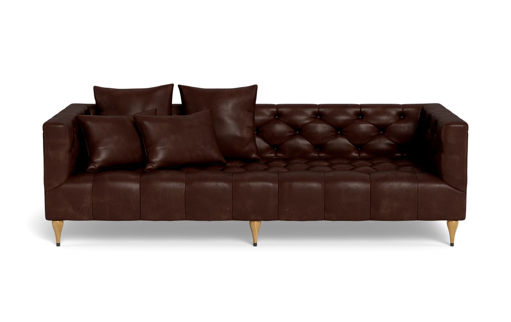 The Ms. Chesterfield Leather Custom Sofa, featuring a luxurious brown leather tufted design and wooden legs, is positioned against a plain background. It is elegantly adorned with four matching cushions, presenting a sleek and modern look.