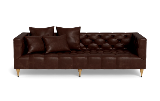 The Ms. Chesterfield Leather Custom Sofa, featuring a luxurious brown leather tufted design and wooden legs, is positioned against a plain background. It is elegantly adorned with four matching cushions, presenting a sleek and modern look.