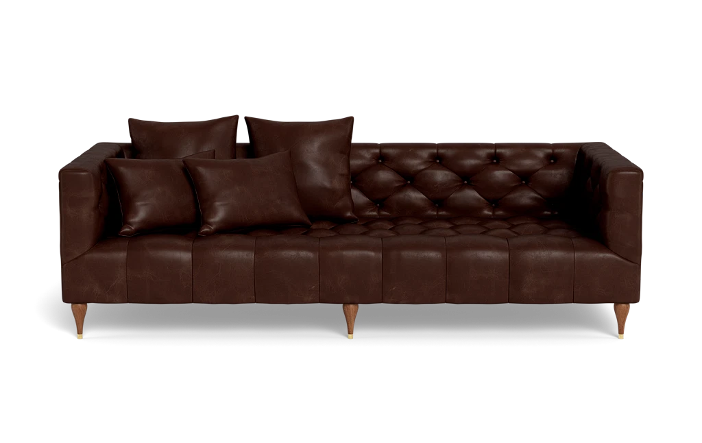 The Ms. Chesterfield Leather Custom Sofa is a dark brown leather piece featuring tufted cushions and paired with four matching pillows. It stands on short wooden legs and is showcased against a plain black background.