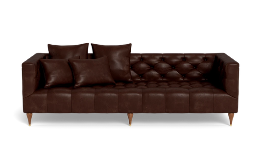 The Ms. Chesterfield Leather Custom Sofa is a dark brown leather piece featuring tufted cushions and paired with four matching pillows. It stands on short wooden legs and is showcased against a plain black background.