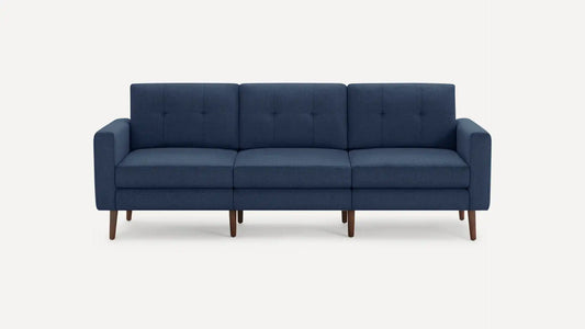 The Nomad Fabric Sofa is a modern, three-seater dark blue piece with a minimalist design, featuring wooden legs. It has tufted back cushions and straight armrests and is displayed against a plain white background.