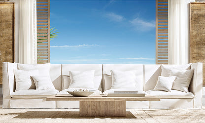 A modern living room featuring a sizable Original Lancaster Leather Recliner adorned with multiple white cushions. In front, there's a wooden coffee table holding a bowl and books. The background presents open windows with slatted wooden panels framing a picturesque view of the blue sky dotted with clouds.