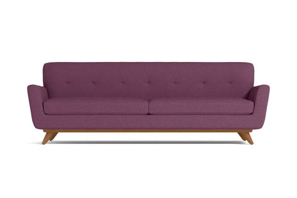 The Carson Sofa is a mid-century modern piece with purple upholstered fabric, featuring a tufted backrest and wooden legs. It boasts a minimalist design and comfortably seats three people.