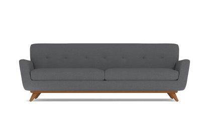 The Carson Sofa offers a modern aesthetic with its dark grey hue and minimalist design. It features a cushioned back adorned with subtle button detailing, sleek armrests, and is supported by mid-century styled wooden legs.