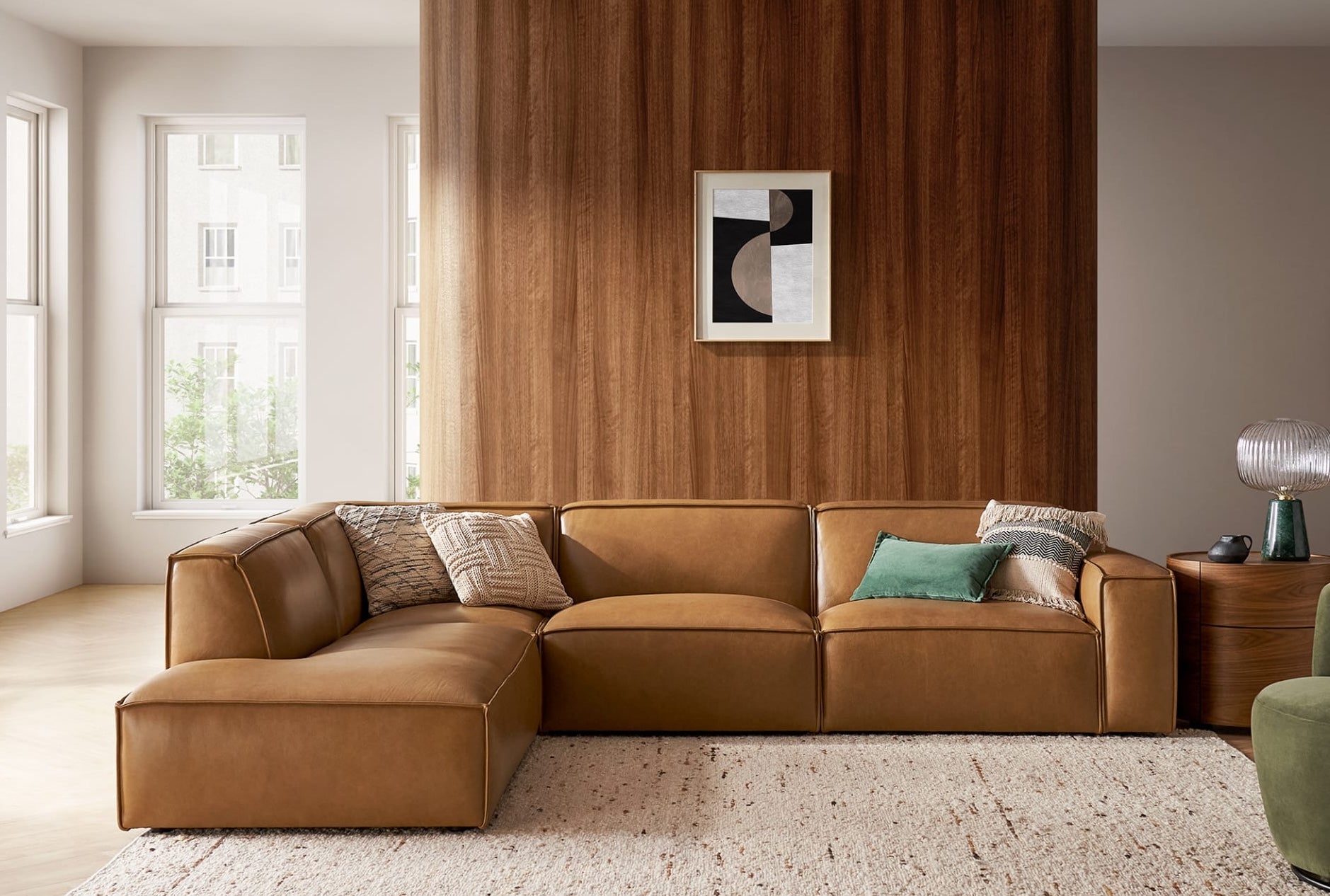 A modern living room with a brown leather sectional sofa adorned with assorted pillows in beige, green, and brown. The backdrop boasts a wooden panel wall with abstract art. Sunlight streams in from large windows, illuminating the beige rug and light wood floors.