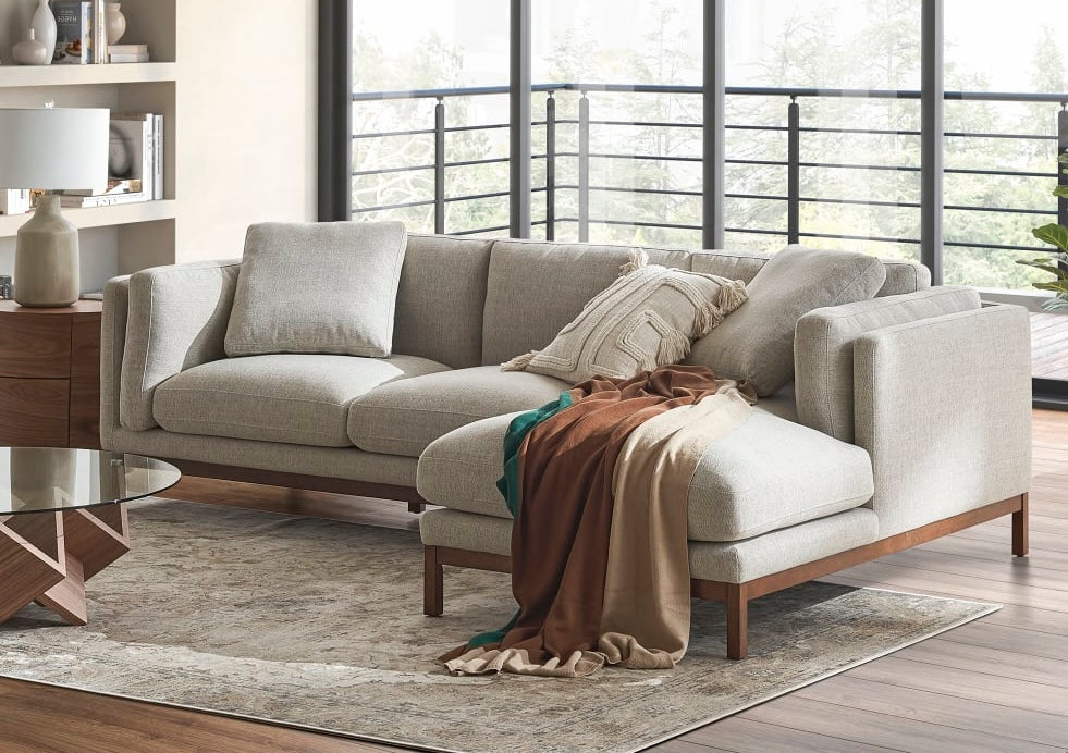 A modern living room features an L-shaped, light gray sectional sofa adorned with assorted throw pillows and a brown and beige blanket. The room includes a glass coffee table on a beige rug and large windows showcasing a view of trees and a balcony.