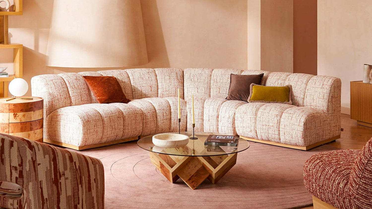 A warmly lit living room featuring a peach-colored, textured sectional sofa with various pillows, including a brown and a mustard yellow one. A round glass coffee table with a geometric wooden base sits on a circular area rug. Shelves and a lamp are in the background.