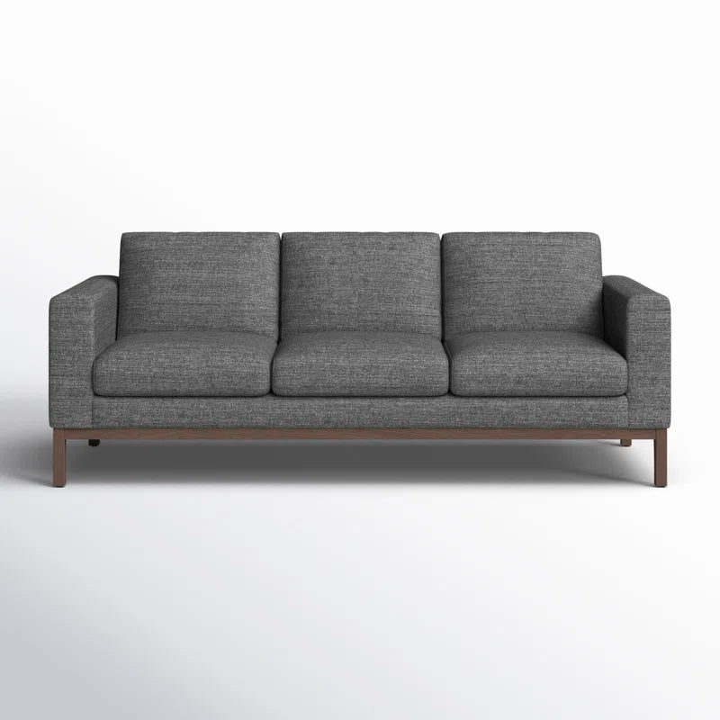 The Clayton 83.1" Upholstered Sofa, a modern piece in gray fabric with three cushions, is displayed against a plain white background. It showcases a clean, minimalist design with wooden legs.