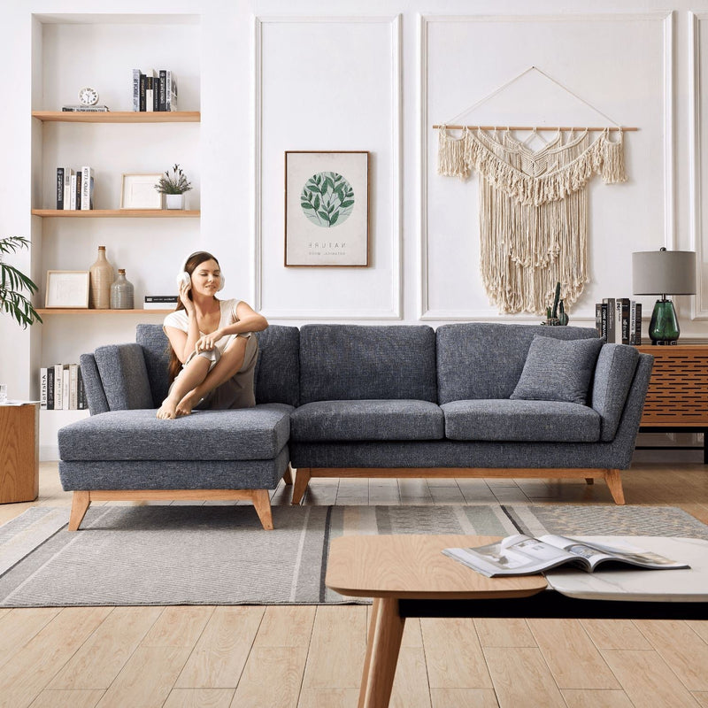 A person sits on a ValMinimal Sectional, a modern gray sofa, in a stylish living room. The room showcases a macrame wall hanging, bookshelves, plants, and a wooden cabinet. A coffee table with an open book is placed in the foreground on a patterned rug.