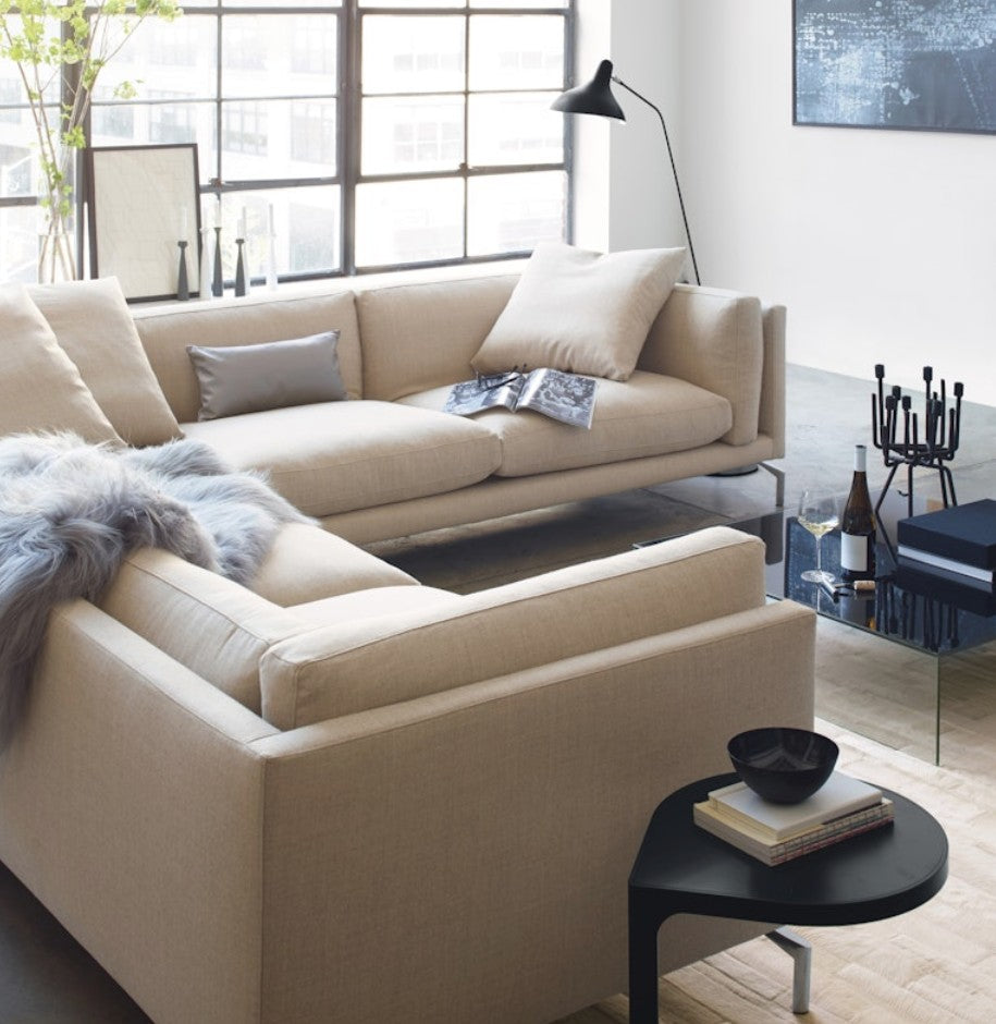 A modern living room featuring a spacious Como Sectional Sofa in beige, adorned with cushions and a gray throw. A sleek floor lamp stands beside it. A small table displays books and decor, while large windows provide ample natural light.