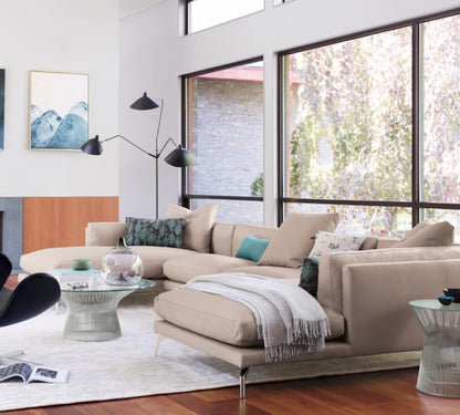 A modern living room with large windows, featuring a Como Sectional Sofa in beige, a glass coffee table, and a stylish floor lamp. The room is adorned with cushions, a throw blanket, and wall art, with trees visible outside the windows.