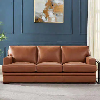 A brown leather three-seat sofa is placed against a dark blue paneled wall. Above the sofa hangs an abstract painting in shades of white, beige, and orange. A beige rug covers the floor and a small side table is visible on the right. A plant is partially seen on the left.