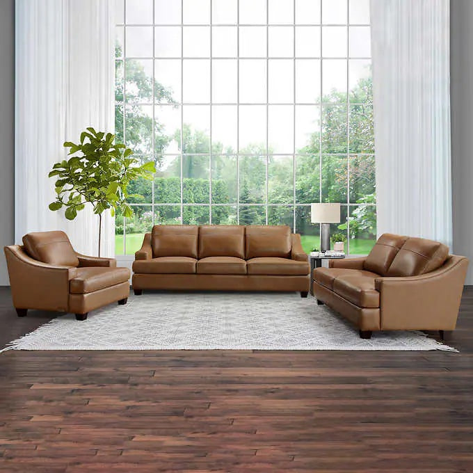 A living room features a brown leather sofa set arranged on a light gray area rug. Large floor-to-ceiling windows with sheer white curtains allow natural light to flood the space. A leafy green plant, lamp, and outdoor view add to the tranquil ambiance.