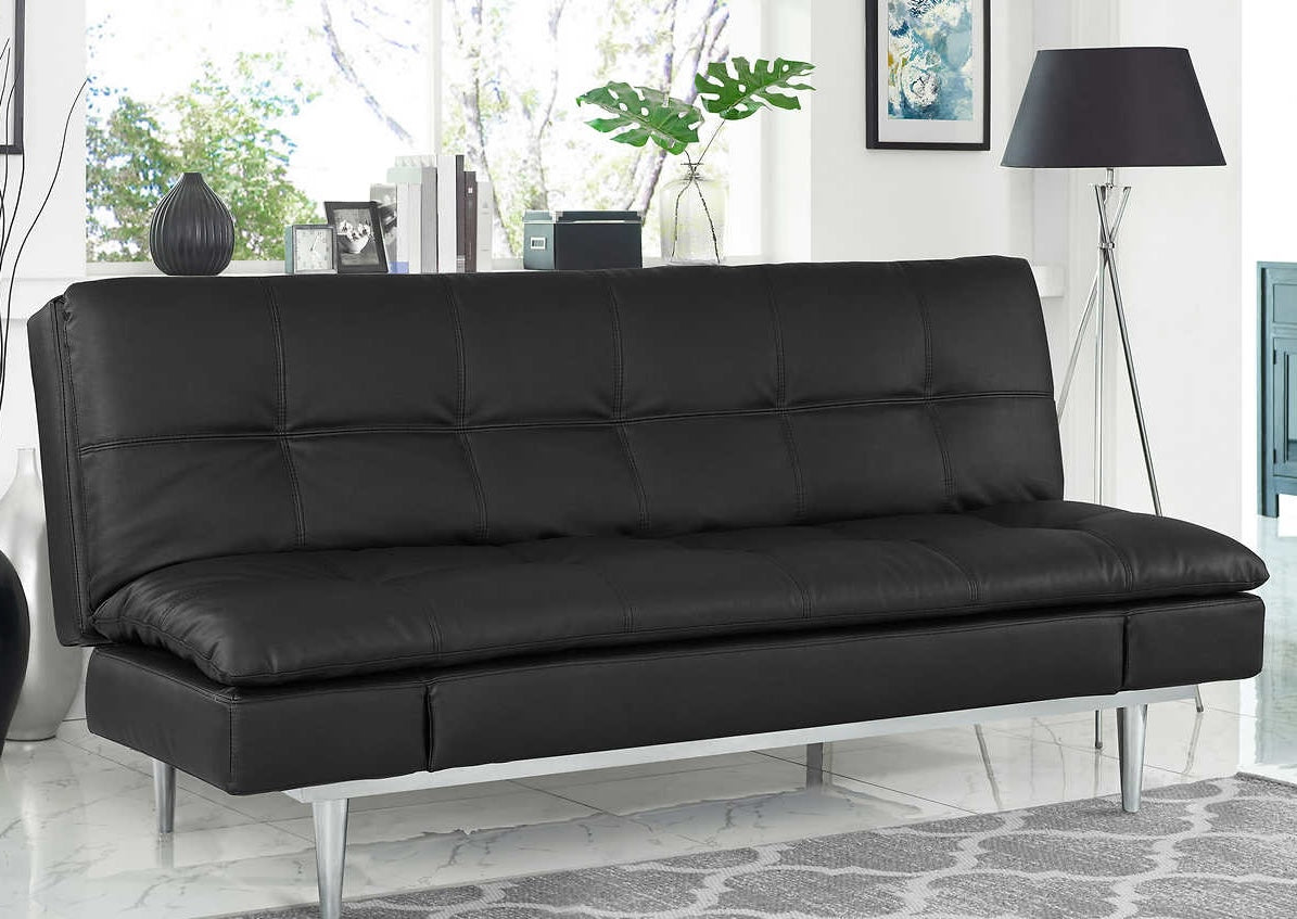 A modern living room features a black faux leather futon sofa with tufted cushions. Behind the sofa, a large window allows natural light to fill the space. The room is decorated with a black floor lamp, green plants, and framed pictures on the wall.