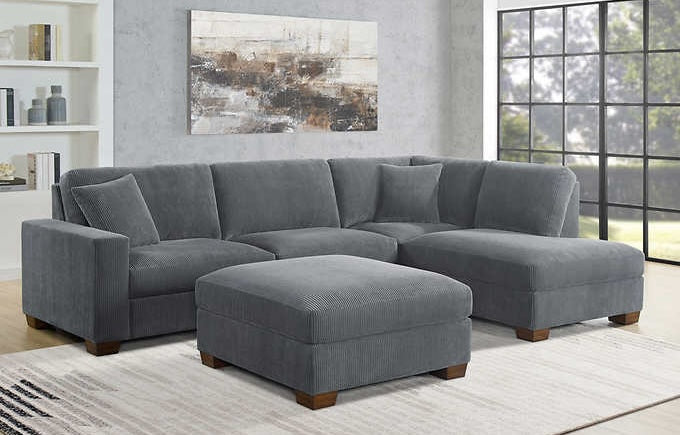 A spacious living room features a modular gray sectional sofa with an ottoman, accessorized with a single matching cushion. The room includes a large window, minimalistic white shelves, a textured rug, and abstract wall art with neutral tones.