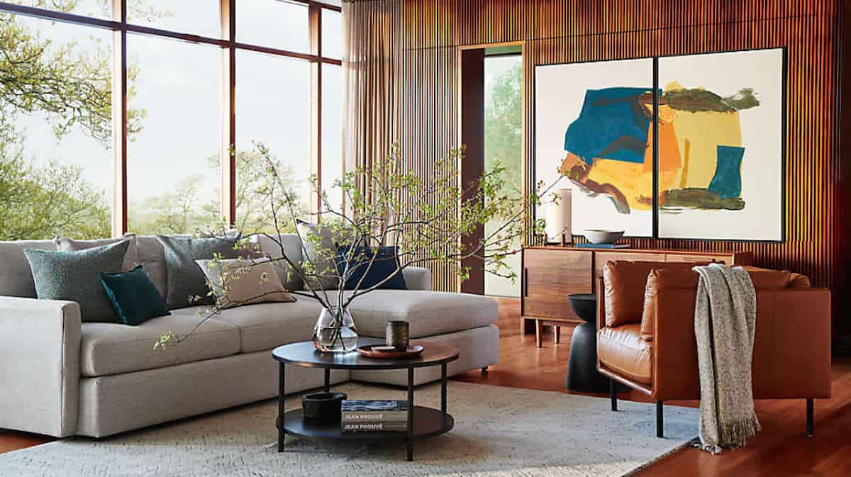 A modern living room featuring a large, light gray sectional sofa with multiple blue and gray pillows. A brown leather armchair with a draped gray blanket sits nearby. A round coffee table with plants and decor is in the center, and there is abstract artwork on the wall.