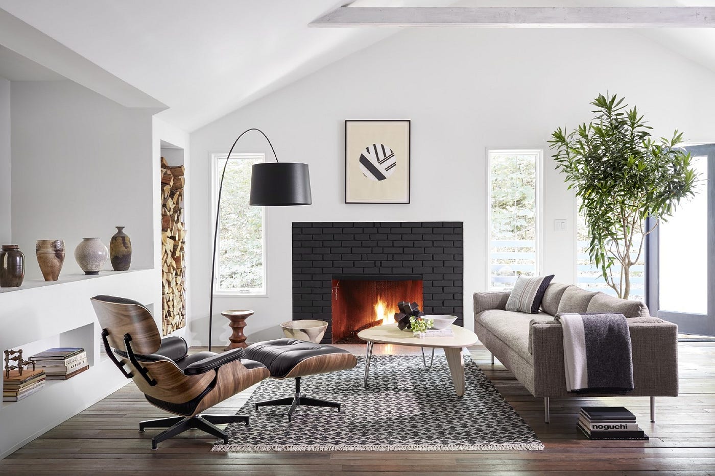 A modern living room features a gray sofa, a wooden lounge chair with an ottoman, a black floor lamp, and a geometric-patterned rug. A black brick fireplace with a fire burning and minimalist art above it accentuates the room. Large windows and a potted plant add brightness.