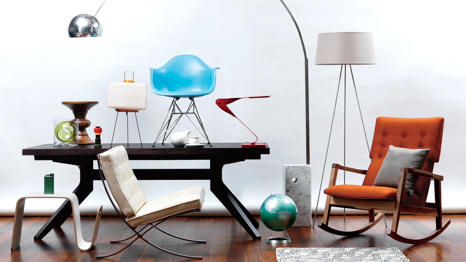 A modern interior displays a variety of furniture and decor items. The setup includes an orange cushioned rocking chair, a white chair, a blue chair, table lamps, a globe, and various decorative objects arranged around a dark wooden table. The floor is wooden and the background is white.